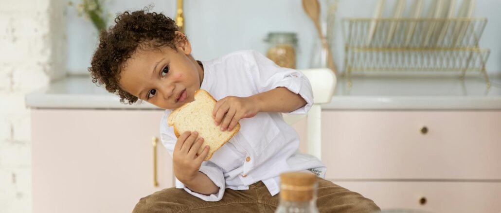 A young boy sits in a cozy kitchen enjoying a slice of bread, surrounded by breakfast items.