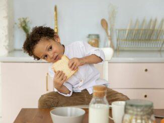 A young boy sits in a cozy kitchen enjoying a slice of bread, surrounded by breakfast items.