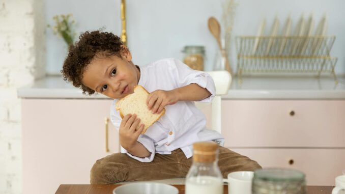 A young boy sits in a cozy kitchen enjoying a slice of bread, surrounded by breakfast items.